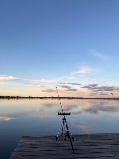 The image depicts a fishing scene at what appears to be a pier or dock extending into a body of water, possibly a lake or river, at sunset. The sky transitions from a light blue overhead to a pale orange and pink near the horizon, reflected in the calm water. Two fishing rods are set up on a rod rest placed on the wooden planks of the dock. There are no people visible in the image. The environment suggests a tranquil, natural setting, likely near Khryashchevka, Russia given the location context.