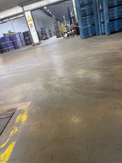 The image shows the interior of a warehouse, likely a distribution center or storage facility, located in Benifairó de la Valldigna, Spain. The floor is made of concrete and has yellow painted markings for safety or lane delineation. Large stacks of blue plastic crates are visible, some filled with products and others empty. In the background, there are loading docks and industrial equipment, including a forklift. The lighting appears to be artificial, typical of an indoor commercial space. The overall environment suggests an active operational facility for goods storage and movement.