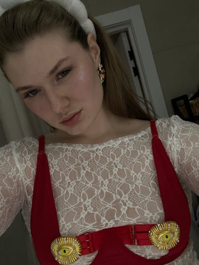 A woman in a white lace top and red suspender-like garment with gold eye-shaped decorations. She is fair-skinned with blonde hair, and wearing an earring on her right ear. The background features a white door and a dark-colored object.