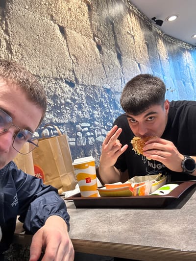 Two males are seated at a table inside a McDonald's restaurant in Krems an der Donau, Austria. The background wall features a large mural depicting a textured stone structure transitioning to a blue sky or water effect on the right. Overhead, recessed circular lights illuminate the area.
The male on the left, partially visible in the foreground from the nose upwards, has short, light brown hair and wears clear-framed eyeglasses. He is dressed in a dark blue windbreaker jacket with a partial "The North Face" logo visible on the left chest. His right hand rests on the table's edge.
The male on the right, visible from the chest up, has short, dark hair with a fade. He wears a black t-shirt with "HUGO" partially legible in white text. A black digital wristwatch is present on his left wrist. He is in the act of consuming a large hamburger, holding it with both hands, his mouth open around the food. Simultaneously, his left hand forms a gesture resembling a "rock on" or "shaka" sign. Both individuals are looking towards the camera.
On the table, a brown McDonald's serving tray holds a partially consumed fast-food meal. Objects on the tray include a yellow and white McDonald's paper beverage cup, the hamburger being eaten by the right male, and several open paper food containers, some appearing empty or containing food remnants. Behind the beverage cup, a brown paper bag with a red logo is visible.
