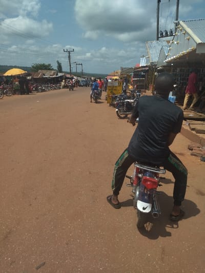 The scene takes place on a dirt road in Lokoja, Nigeria, at latitude 7.404 and longitude 7.632. It is a bright day with a few clouds in the sky. There are several people in the scene, including a man riding a motorcycle. There are also several motorcycles parked on the side of the road, along with a few shops and buildings. The man on the motorcycle is wearing a black shirt and pants, and is looking forward. He is riding a blue motorcycle with a white Nike swoosh on the back fender. The road is made of dirt and gravel, and there are power lines running overhead. There are also some trees and bushes on the side of the road.