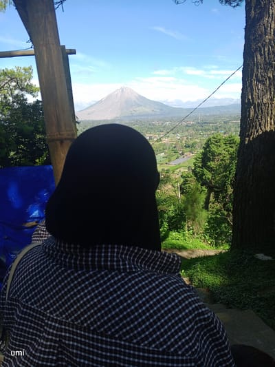 In the city of Gundaling 2, Indonesia, a person wearing a black hijab and a checkered shirt is seen from behind, looking out at a panoramic view of a valley and a distant volcano under a clear blue sky with scattered clouds. The person is positioned on a slightly elevated area, with a large tree trunk to their right and bamboo structures to their left. The foreground includes a blue tarp and dense green vegetation. The valley below shows a patchwork of fields and buildings, suggesting a rural or semi-urban landscape. The volcanic mountain, with its conical shape and visible slopes, is the dominant feature in the background. The lighting suggests it is daytime, with sunlight casting shadows on the ground and vegetation. A watermark "umi" is visible in the bottom left corner of the image.