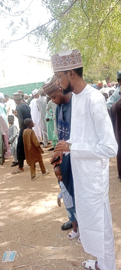 The image depicts an outdoor gathering in Sani Mai Nagge, Nigeria, under natural daylight. In the foreground, a man with a beard, wearing a white patterned kufi cap and a long-sleeved white kaftan with subtle vertical patterns, stands looking left. His right hand holds a dark mobile phone, partially obscured. He wears white open-toed sandals. To his immediate left and slightly behind, another man with a patterned kufi cap wears a dark blue kaftan with light blue geometric embroidery, looking downwards. A small child, wearing a light blue striped shirt and denim shorts with pink sandals, is partially visible peeking from behind the man in the blue kaftan.
The environment consists of light brown, unpaved ground, scattered with small pieces of debris, including wood fragments and what appears to be a discarded piece of light blue material. In the background, numerous individuals, primarily men and boys dressed in various traditional tunics (white, light blue, brown, black) and caps, are gathered. A light-colored wall with some discolored patches is visible on the left side of the background. Green foliage from a tree is present in the upper right portion of the image. Shadows indicate direct sunlight. No specific interactions beyond general gathering are discernible.