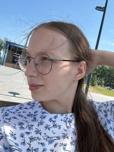A young woman with light brown hair, wearing round glasses and a white dress with a blue floral pattern, is captured in a medium close-up shot. Her hair is slightly windswept, with strands blowing across her face and head. She is looking to her left, with her right hand resting on the back of her head. The background suggests an outdoor urban environment, possibly a park or promenade, with paved ground, some trees, a lamppost, and a building with text on it. The sky is clear and blue. The image appears to be taken outdoors on a sunny day.