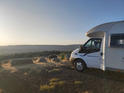 A white recreational vehicle, identified by the word "TRANSIT" on its side, is parked on a gravel surface overlooking a vast, tree-covered landscape. The RV is positioned on the right side of the frame, facing left, with its front wheel visible and the driver's side door slightly ajar. The landscape stretches out towards a hazy horizon, with rolling hills and scattered vegetation under a clear, pale blue sky. The foreground features several large, weathered rocks scattered on the gravelly ground, which is sparsely covered with dry grass. The scene appears to be set during either sunrise or sunset, with warm, soft light illuminating the landscape and casting long shadows. The location is identified as being in the city of Ledrada, Spain.
