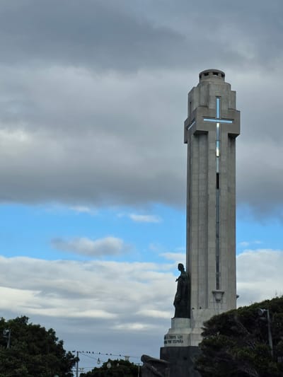 This image shows the Monument to the Battle of the Little Big Horn, also known as the Cross of the Canaries, located in Santa Cruz de Tenerife, Spain. The monument is a tall, concrete tower with a cross at the top, which is lit with blue lights. At the base of the tower is a statue of a person. The monument is surrounded by trees and a cloudy sky.