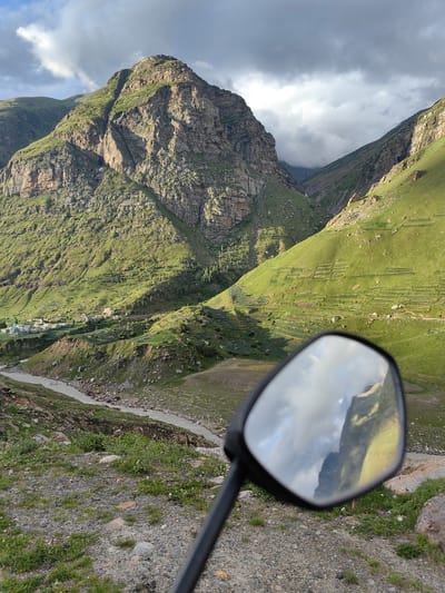 A motorbike's side mirror reflects a dramatic mountain landscape in Yari-Khoksar, India. The mirror shows a jagged, sun-drenched mountain peak with green slopes and a cloudy sky. In the foreground of the main image, the mirror is attached to a black handle, positioned over a rocky, gravelly terrain dotted with sparse green vegetation and small purple flowers. Beyond the mirror, a winding, light-colored river flows through a valley. The valley walls are steep and covered in lush green grass, with evidence of terraced farming on the slopes. A small cluster of buildings, likely a village, is visible in the mid-ground on the left side of the valley. The sky above is a mix of dark, brooding clouds and patches of bright sunlight, casting shadows across the mountains. The overall scene depicts a remote and rugged natural environment.