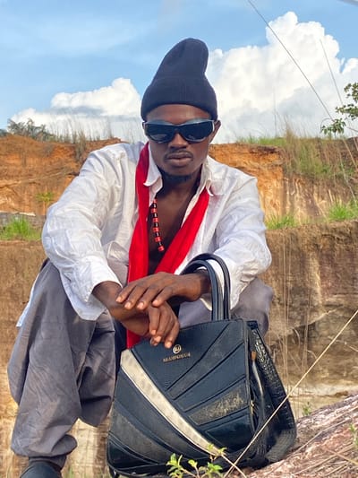 An outdoor portrait shot features an individual sitting or crouching in a natural, rugged environment, looking directly at the camera. The subject is an adult male, wearing a black beanie, dark sunglasses, a white long-sleeved shirt, and dark pants. A bright red fabric is draped around his neck, under which a black and red beaded necklace is visible. His hands are clasped in front of him.
Beside him, resting on the ground, is a black handbag or briefcase with a geometric design, silver-colored accents, and a light-colored lining. The brand name "BRAND OMONO" is faintly visible in metallic letters on the bag.
The background consists of layered rock formations or cliffs, indicating an exposed geological area or a quarry. Sparse green grass and dry vegetation are scattered across the rocky terrain. The sky above is bright blue with prominent white cumulus clouds, suggesting it is daytime with clear weather. The setting appears to be a natural landscape in Afikpo, Nigeria. The subject's pose implies he is intentionally posing for the photograph, presenting a cool and composed demeanor.