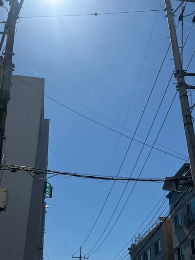 The image captures a worm's-eye view of a bustling urban sky in Cheongju-si, South Korea, dominated by a complex network of utility infrastructure against a clear, bright blue sky. The sun is visible in the lower-left corner, casting a strong lens flare and illuminating the scene.
Two large utility poles or crossarms span the frame. One stretches across the top, while another is situated lower down, appearing to have electrical equipment attached, where faint numbers and letters, possibly "900 20," are visible. A dense web of black electrical and communication wires crisscrosses the sky, connecting these poles and extending towards buildings. A black cable or rope with a metallic loop hangs vertically from the top utility structure on the left.
Parts of two distinct buildings are visible. In the upper-right quadrant, a multi-storied building with numerous windows and a section of brick facade is seen from below. To the middle and lower right, the roofline and upper section of a larger, light-colored building are present. This building features a flat roof with various structures, including a prominent green, arched skylight or shelter, and other small pieces of equipment.
The scene is static, showing no human activity. The bright sun and clear sky indicate a sunny daytime environment, likely mid-morning or afternoon. The dense overhead wiring and modern building architecture are characteristic of an urban setting.