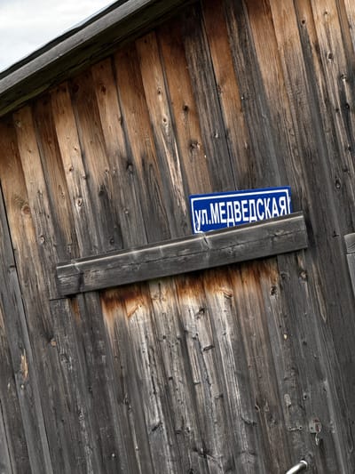 The image shows a blue and white street sign reading "ул. МЕДВЕДСКАЯ" (ul. MEDVEDSKAYA) vertically mounted on a weathered wooden wall or structure. The sign is partially obscured by a gray wooden support beam. The wooden planks of the wall are gray and brown with visible knots and grain. A metal object (possibly a lock or handle) is visible in the upper right corner of the image. The scene likely depicts a location on Medvedskaya Street, in a place named Medvedka.