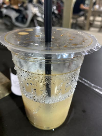 The media file presents a close-up view of a clear plastic cup containing a light-colored, possibly milky, beverage. The image is rotated 90 degrees clockwise, making the cup appear to be resting on its side on a dark, reflective surface. A black straw is inserted through the clear plastic lid into the drink.

A prominent feature is the heavy condensation on both the exterior and interior of the cup, manifested as numerous small water droplets, indicating the beverage is cold. Some darker, possibly brown, liquid residue or drips are visible on the underside of the plastic lid, near where it seals the cup. The beverage itself appears to be a uniform light beige or yellow hue.

The background is out of focus, showing blurred shapes and highlights, suggesting an indoor setting like a cafe, restaurant, or perhaps a casual home environment. No specific objects or details in the background offer further clues about the location within Gampongaceh, Indonesia, or the time of day. No text is visible on the cup, straw, or in the surrounding area. The image primarily focuses on the textured details of the cold drink container.