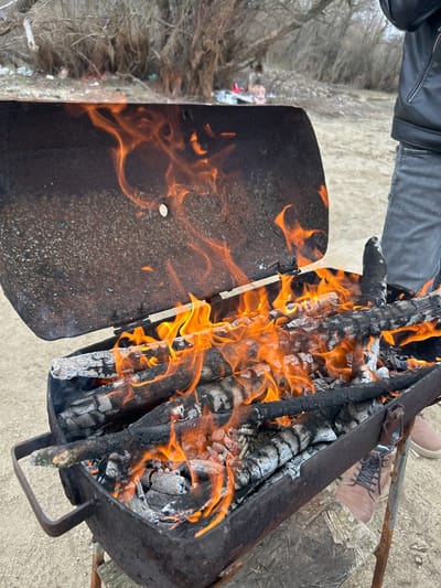 A portable, rusted metal grill with its hinged lid open is actively burning wood logs and embers outdoors. Bright orange flames emanate from the charred wood within the rectangular basin. The grill's interior surfaces are dark with soot and exhibit signs of corrosion. In the lower right of the frame, a person stands, visible from the torso down, wearing a dark leather jacket, faded blue jeans, and brown lace-up shoes. The scene is set on light-colored, sandy ground, with leafless trees and dense bushes occupying the blurred background. Indistinct colorful items, consistent with refuse, are visible near the base of the distant trees. This event is located in the city of Любен, Bulgaria.