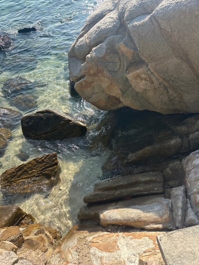 The image depicts a rocky coastline scene in ตำบล เกาะเต่า, Thailand, under bright daylight. The foreground and midground feature clear, shallow ocean water with sunlight reflecting off its surface, creating shimmering patterns. Submerged beneath the transparent water, a light-colored sandy seafloor is visible along with several dark-brown and grey rocks of varying sizes.
To the upper right and extending into the center, a massive, light grey-brown boulder with visible fissures and textured surfaces dominates, partially submerged in the water. Below this large boulder, the water appears darker due to shadow and depth. Smaller, layered rock formations, brownish and grey, line the immediate foreground and extend into the water, forming the irregular edge of the landmass. No people, animals, or man-made objects are present in the scene. The overall environment is a natural, undisturbed coastal rock formation interacting with calm, clear seawater.