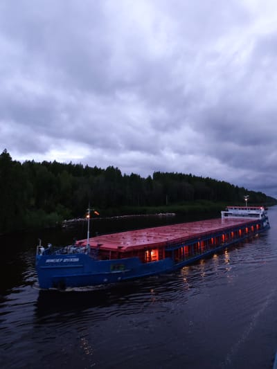 The image shows a large, blue and red barge traveling down a canal or river. The barge is lit from within, with orange lights visible. The sky is overcast with dark clouds, suggesting it is either dusk or dawn. Lush green trees line the banks of the waterway. The bow of the vessel has writing on it in Russian, with the name "инженер Шиткова" clearly visible, suggesting its location near Aleksandrovskoe, Russia. The water is dark and reflects the dim light.