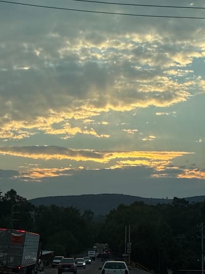 The image depicts a view along a road in Santiago Tlautla, Mexico, at dusk or dawn. The sky is filled with clouds, some illuminated by golden sunlight. A dark, forested mountain range is visible in the distance. In the foreground, a line of traffic, including cars and a large truck, is observed. Electrical wires are visible in the sky.