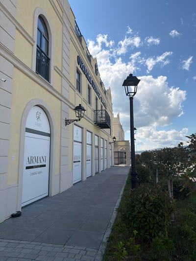 The image captures a storefront exterior on a sunny day in Gamarjveba, Georgia. The building, painted a pale yellow, features arched windows and decorative molding. Several large white signs on the storefront windows display "ARMANI OUTLET COMING SOON" along with the "NABU" logo. A tall black lamppost stands prominently on the right side of the image, with a smaller one mounted on the building. The walkway in front of the stores is paved with gray tiles. To the right of the walkway, lush green bushes and trees are visible. The sky above is blue with scattered white clouds.