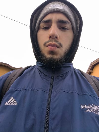 The image is a low-angle, close-up selfie of a male individual in Meistratzheim, France. The subject, with a beard and mustache, looks directly at the camera with a neutral expression and slightly pursed lips. He is wearing a grey knitted beanie featuring a white Nike swoosh logo, a dark-colored hoodie pulled over the beanie, and a dark blue zip-up jacket. The jacket displays a white "adidas" logo on the left chest and a mirrored "Michelob ULTRA" logo on the right chest. Dark backpack straps are visible over his shoulders. The background consists primarily of a bright, overcast sky with a thin dark utility wire crossing horizontally. Portions of a reddish-brown roof structure are visible on the lower left, and an orange-yellow building facade with brown eaves is partially visible on the lower right.