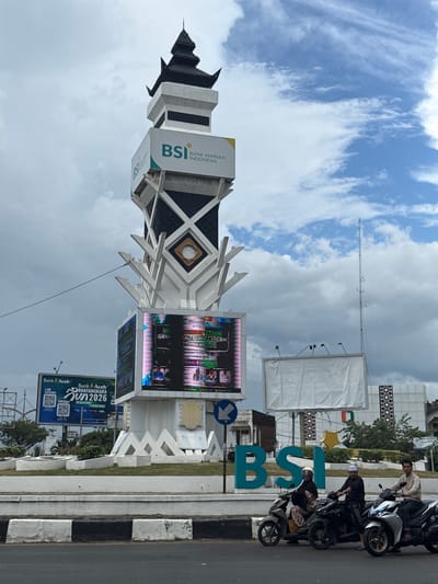 The image captures a street view in Lambaro, Indonesia, dominated by a large, multi-level tower structure. The tower is adorned with signage, including a prominent "BSI BANK SYARIAH INDONESIA" logo on its upper section. A large LED screen on the lower part of the tower displays colorful content, featuring images of people and text. In the foreground, several individuals are riding motorcycles. On the left side of the frame, a billboard advertises "Bank Aceh" and "Run 2026." Behind the tower, a tall communication tower is visible. The sky is partly cloudy, with a mix of blue sky and white clouds. The overall scene suggests a commercial or public area within the city.