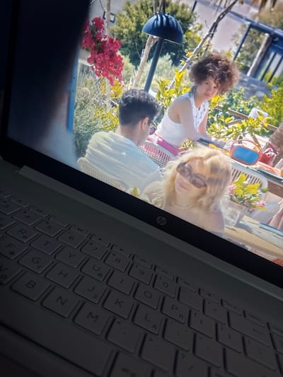 The image shows a laptop screen displaying a scene from a television show or movie. The on-screen content features a group of young adults outdoors, possibly at a cafe or a terrace, with lush green foliage and pink flowers in the background. The individuals appear to be engaged in a conversation or enjoying a meal. The lower portion of the image captures the keyboard of the laptop, with the keys illuminated by the screen's light. The overall setting suggests a casual, sunny day. The image does not provide any information regarding the location in Paralimni, Cyprus, or any specific details that would link it to that city.
