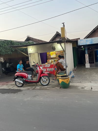 The image depicts a street scene in Karangtanjung, Indonesia, likely during the late afternoon or early evening due to the soft, diffused light. In the foreground, a red and silver scooter is parked on the side of a paved road. Two individuals are visible near the scooter. One, wearing a light blue shirt, is seated on the scooter, and another, wearing dark clothing, is standing behind him.
Behind the scooter is a small roadside stall or shop. A man, wearing a white shirt and a patterned sarong-like garment, is seated on a large, decorative object that resembles a stylized watermelon or gourd, painted green and yellow. He appears to be interacting with the stall. The stall itself has a canopy and displays various items, including what look like packaged goods and a prominent red banner with white text. Visible text on the banner includes "ES DROP," "JAK YAKIS," and "CARI ALL K." Another sign above the stall is yellow and appears to have text on it as well, but it is less legible. The building behind the stall has a corrugated metal roof and white walls, with some decorative elements. Green vines or foliage are visible growing on the side of the building and along a fence. Overhead, electrical wires crisscross the sky, which is a pale, hazy white, suggesting a cloudy or overcast sky. The overall impression is of a casual, everyday scene at a local vendor.