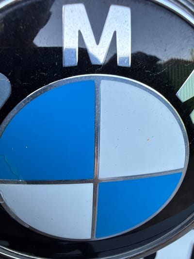 The image shows a close-up of a BMW logo. The logo consists of a circular emblem divided into four quadrants, alternating between blue and white, with the letters "BMW" inscribed above it in a silver, metallic font. The emblem is set against a black background, which appears to be a car's exterior. The image is taken outdoors, with hints of natural light and a faint reflection of what might be foliage or a fence visible in the background. The logo itself appears slightly weathered, with minor scuffs and dust particles on its surface. There are no people or any other identifiable objects in the immediate vicinity of the logo within the frame. The location in the city of Петропавловка, Kyrgyzstan, cannot be determined from the visual information provided in the image.