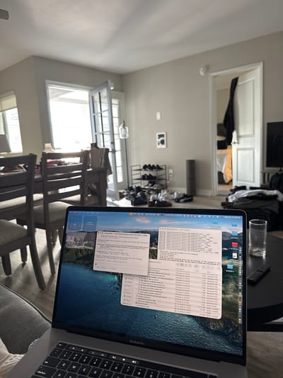 A low-angle, slightly canted indoor shot shows a MacBook Pro displaying a Finder window and a Terminal window, on a gray upholstered seat. The screen shows a list of markdown files in Finder and a command-line interface in Terminal. To the left, through a doorway, is a well-lit interior space with a window that lets in daylight. The house appears modern, with simple paint colors and trim work. In the background, there are indications of home life: clothing, shoes, a table. The scene evokes a sense of working from home in a comfortable, modern residential setting. I cannot confirm the location in the city of Redondo Beach, United States of America.