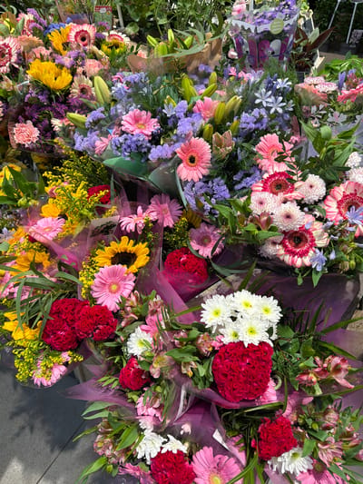 The image shows a close-up, eye-level view of several large bouquets of flowers displayed outdoors, likely at a market or flower shop. The bouquets are composed of a variety of colorful flowers including pink and white gerbera daisies, yellow sunflowers, red cockscomb celosia, purple statice, and yellow solidago. The flowers are wrapped in translucent purple paper and are densely packed together. The background is blurred, but appears to be a mix of greenery and more flowers, suggesting a vibrant floral display. The ground beneath the bouquets is visible in the lower left corner and appears to be a gray paved surface, possibly a sidewalk or patio. There are no people or animals visible in the image, and the focus is entirely on the floral arrangements. The lighting suggests it is daytime, with natural light illuminating the scene.