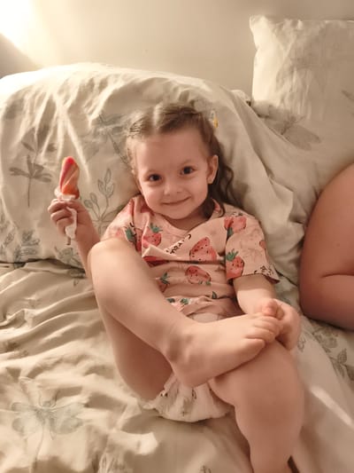 A young girl with brown hair tied in pigtails is lying on a bed covered with a patterned duvet. She is wearing a pink t-shirt with a strawberry pattern and a diaper. She is holding a multi-colored popsicle in her right hand and smiling at the camera. Her legs are crossed, with her right leg on top of her left. The background is a plain wall with some lighting creating shadows. The overall mood is relaxed and happy. The visible text on the t-shirt reads "merci". The scene appears to be indoors, possibly in a bedroom, and the time of day is likely evening or night due to the lighting.