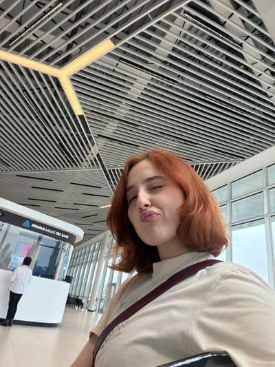 The photo is taken inside the David the Builder Kutaisi International Airport in Ianeti, Georgia. A young, red-haired woman is oriented top-left of the frame, looking at the camera with pursed lips. She is wearing a light-colored shirt with a burgundy stripe on the sleeves. To her left is a large metal grid with LED lighting. Below her to the right is a TBC Bank kiosk with a person dressed in white and black standing at the counter. The kiosk appears to be white with reflective surfaces. Other travelers are visible further in the background. Above the travelers is a window looking out to the sky. The photo is taken at a sharp angle.