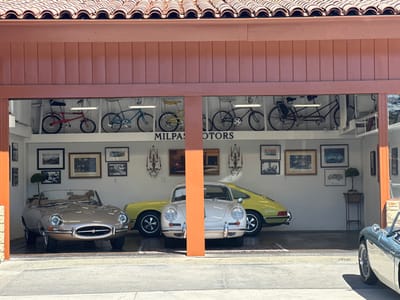 An open-fronted showroom or garage, located in Montecito, United States, is visible under bright daylight. The building features a terracotta barrel-tiled roof and an orange-brown painted fascia. Inside, the white-walled interior displays classic vehicles and bicycles.

On the ground level, a tan Jaguar E-Type Series 1 roadster is centrally parked, facing forward. To its right, a light yellow early-model Porsche 911 is partially visible, parked sideways, alongside a light beige Porsche 356 coupe facing forward. The floor is light gray concrete. Approximately a dozen framed pictures and artworks, along with two ornate dark metal wall sconces, adorn the walls. A small, conical topiary plant in a dark pot is positioned on a stand near the right wall.

Above, on a white shelf, five classic bicycles are displayed: a red BMX-style, a blue cruiser, a yellow child's bike, a black men's bicycle, and a black tandem. Linear lighting illuminates this display. The words "MILPAS MOTORS" are painted in black on the wall beneath the bicycles.

Outside the structure, on the far right, the front-right fender and wire wheel of a light blue/green classic British roadster, consistent with an Austin-Healey 3000, are partially visible. No individuals are present.