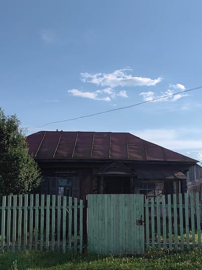 A rustic, old wooden house with a dark, weathered appearance stands behind a pale green, picket fence. The house has a prominent, sloped roof covered in what appears to be dark metal, possibly corrugated iron, with visible seams running diagonally. The house itself is dark brown, with visible wooden siding. A small, ornate porch structure with decorative trim is visible above the entrance. One window to the left of the porch is visible, with dark panes. The fence in the foreground is constructed of vertical wooden planks, painted a faded green, and features a gate on the left side secured with a simple latch. Beyond the fence, the ground is covered in green grass and some sparse vegetation. To the left of the house, a large, leafy green tree is partially visible. The sky above is a bright blue, with a few scattered white clouds. The scene suggests a rural or semi-rural setting, likely in a village or small town. The image was taken in the city of Krestovo-Gorodishche, Russia.