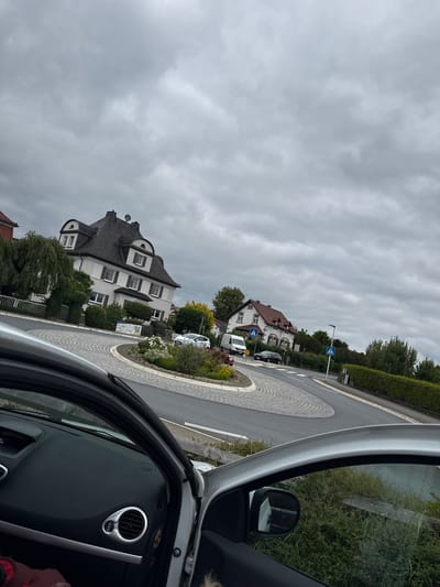 The image depicts a view from inside a car looking out at a roundabout in Braunfels, Germany. The sky is overcast with gray clouds. On the left side of the roundabout, a large white house with a dark, steep roof and dormer windows is visible, surrounded by greenery and a fence. A white car is parked on the roundabout. Further along the road, another house with a red-tiled roof and a black car is seen. Pedestrian crossing signs are present on the roadside. The car interior, including the dashboard and door frame, is visible in the foreground. The road surface of the roundabout is paved with cobblestones in a circular pattern. The overall impression is an ordinary urban street scene on a cloudy day.