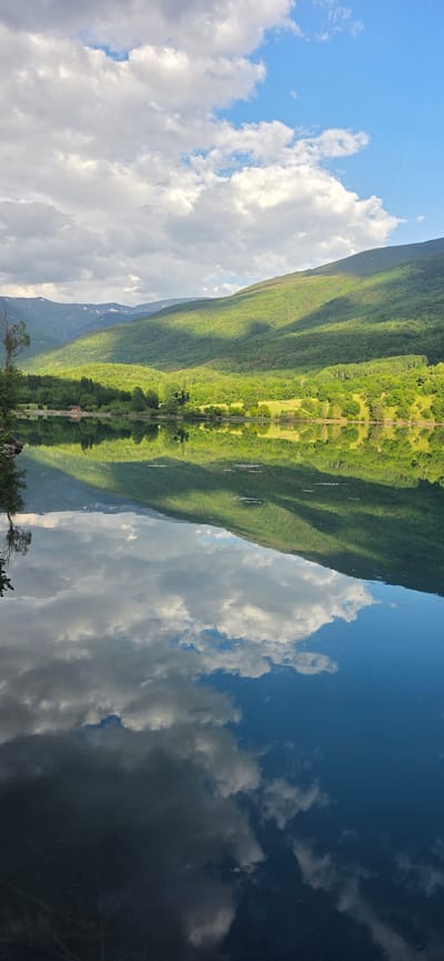 This is a vertical, eye-level, medium shot of a serene lake reflecting a verdant, tree-covered mountain range under a sky with scattered white clouds. The image captures the natural beauty of the landscape near Sahún, Spain, possibly the Embalse de La Tranquera, a reservoir that reflects the surrounding scenery. The foreground is dominated by the still water of the lake, which perfectly mirrors the sky and the lush green slopes of the mountains. The mountains themselves are dappled with sunlight and shadow, indicating a dynamic interplay of light and shade. Along the far shore, a dense forest meets the water's edge, and a small, reddish building is visible amongst the trees. The sky above is a bright blue, interspersed with fluffy white clouds, some of which are illuminated by sunlight. The reflection in the water is so clear that it's difficult to distinguish from the actual scene, creating a symmetrical and tranquil visual experience. There are no people or animals visible in the image, emphasizing the quiet and untouched nature of the location.
