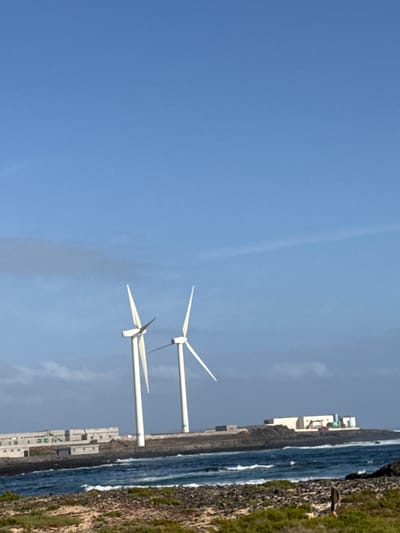 An outdoor image, originally rotated, depicts a clear, sunny coastal scene in Corralejo, Spain. The foreground is dominated by a vast expanse of clear blue sky, with only subtle atmospheric haze visible in places.
In the mid-ground, two large, white wind turbines stand prominently. Each turbine features a tall, slender mast and three elongated blades, characteristic of modern wind energy generators. They appear active or ready for operation.
Below the wind turbines, the scene transitions to a coastline. The calm, blue-green sea extends across the middle and right side of the frame, with gentle white waves breaking near the shore. Along the immediate coast, there's a strip of dark, rocky terrain, which then gives way to patches of green grass and vegetation further inland on the far right.
Several white, multi-story buildings are situated along the coastline, suggesting a resort, residential area, or coastal town. Some buildings have flat roofs and appear to be of a contemporary architectural style; one building shows green detailing.
On the far right, near the edge of the land and grassy area, a single person is visible, standing and seemingly observing the sea or the surroundings. They are wearing dark clothing.
The time of day appears to be bright daylight, possibly midday or early afternoon, indicated by the clear sky and strong illumination. The weather is clear and calm. There is no visible text in the image.