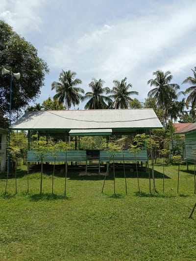 The image shows a light blue, stilted structure with a corrugated metal roof, located in Blangkarieng, Indonesia. The structure is surrounded by a low fence of thin wooden stakes. The foreground is a green lawn. In the background, there are palm trees and other buildings. A loudspeaker is mounted on a pole to the left of the structure. The sky is partly cloudy.