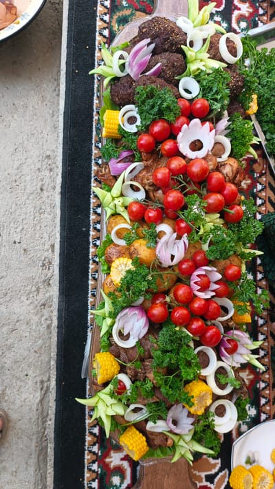 A top-down, close-up, high-angle shot captures a large platter of food, likely a traditional Uzbek meal, placed on a rug. The platter is loaded with grilled meats, including what appear to be kofta and chicken pieces, interspersed with roasted potatoes, corn on the cob segments, cherry tomatoes, red onion slices, and rings of white onion. Fresh parsley sprigs are used as a garnish throughout. The food is arranged in a decorative manner, with some onion slices carved to resemble flowers and cucumber slices artfully placed as accents. The setting appears to be outdoors, on a concrete surface beside a traditional rug. The overall impression is of a festive and abundant meal.