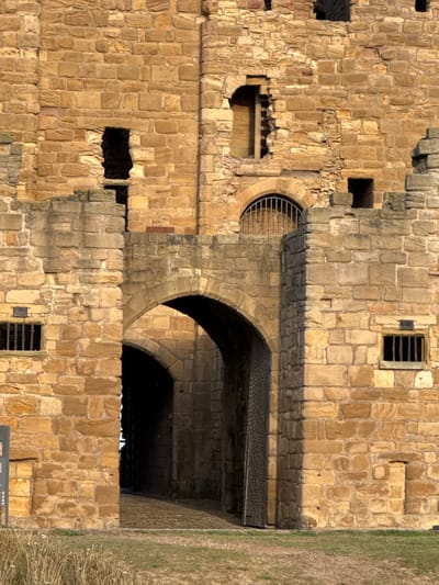 The image displays a detailed view of the stone architecture of Tynemouth Priory and Castle, located in North Tyneside, United Kingdom. The scene is dominated by extensive walls constructed from rough-hewn, light brown and grey sandstone blocks, showing evidence of significant weathering and repair.
In the lower central area, two prominent arched passageways are visible. The foremost archway is deep and dark, revealing a paved path extending inward. To the right of this primary arch, a heavy, vertically planked wooden door or gate is partially open, leaning against the inner wall of the passage. A secondary, smaller arch is visible deeper within and to the left of the main opening.
Above these arches, the walls continue upward, featuring several irregular openings and recesses. A higher arched opening with a metal grate is positioned above the main archway. To the left, a large, irregular opening in the upper wall reveals what appears to be a portion of an internal stone staircase. Two smaller, rectangular openings fitted with vertical metal bars are situated on the lower parts of the left and right walls. The overall structure exhibits battlements and eroded sections along its upper edge.
The scene is illuminated by direct sunlight, casting distinct shadows that highlight the texture of the stone and create deep contrast within the archways and recesses. The ground in the immediate foreground consists of dry, patchy grass and sparse vegetation. No individuals, animals, or active subjects are present in the frame.