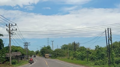 The image depicts a two-lane road in Paku, Indonesia, flanked by lush green vegetation and power lines. In the foreground, a person on a motorcycle, wearing a red jacket and a black backpack, is riding away from the camera. Further down the road, a yellow truck is visible. Utility poles with multiple wires are prominently featured throughout the scene, stretching across the sky, which is partly cloudy with a blue sky. A small, rustic structure with a thatched roof is situated to the left of the road, near a utility pole. The scene suggests a rural or semi-rural setting within Paku.