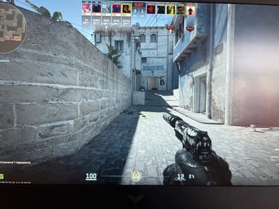 The image is a screenshot from a first-person shooter video game, specifically Counter-Strike: Global Offensive, displayed on a laptop screen. The in-game environment depicts a street scene within a city. The camera perspective is from a player holding a handgun with a distinct black and white patterned skin. The player is positioned on a cobblestone path, looking down a narrow alleyway flanked by stone and light-colored buildings. A minimap is visible in the upper left corner, showing a portion of a game map labeled "A." The upper portion of the screen displays player names, scores, and health/armor information, along with a timer showing "0:07." The bottom of the screen shows game statistics, including ammunition count ("100") and player status. The in-game text also indicates that the current objective is "устранение 1 террориста" which translates to "eliminate 1 terrorist." There is no indication or information within the image that places the scene in Lebedinovka, Kyrgyzstan.