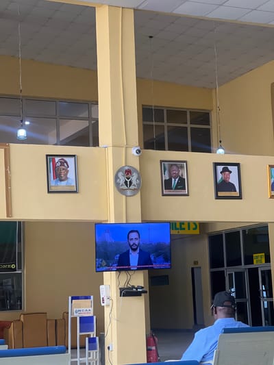 The image shows the interior of a building in an unknown location in Kwi, Nigeria. Displayed on a yellow pillar are framed pictures of political figures, including Festus Keyamo and Bola Ahmed Tinubu, as well as a television displaying news. The Nigerian coat of arms is also displayed on the pillar. A man in a blue shirt and a black hat is partially visible in the upper-right corner of the image. A sign that reads "SECURITY DUTY OFFICE" is visible above the man. A fire extinguisher is mounted near the security guard. Near the television is some shelving with "ONCAA" printed on it. A sign that reads "TOILETS" can be seen above the security guard's head.