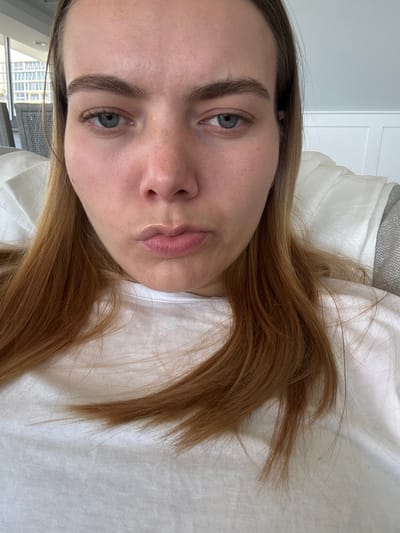 A woman with light skin and light brown hair lies on a white surface, possibly a bed. Her eyes are light blue, and her eyebrows are thick and slightly arched. Her lips are full, and her expression is neutral. A white T-shirt is visible beneath her face. Background includes white wall with door.