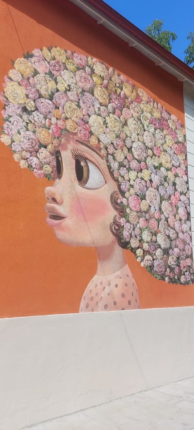 This is a mural painted on an orange wall, depicting a stylized portrait of a person. The person's head is entirely made of a dense arrangement of various pink, yellow, and white flowers, resembling roses. Only a portion of the person's face and neck are visible, emerging from the floral mass. The visible facial features include a large eye with thick eyelashes, a small nose, and lips. The person is wearing a light-colored garment with dark polka dots on the sleeve and neckline. The mural is painted in a bright, somewhat whimsical style.
To the left of the mural, a section of a white-sided building with a red gutter and white eaves is visible against a bright blue sky with some green tree branches. To the right of the mural, a different wall section is visible, painted a light off-white color. A thin, vertical line divides this wall from another section that appears to have a subtle brick or tile pattern.
The lighting suggests it is daytime, with the sun casting shadows from the building's eaves onto the orange wall. The overall impression is cheerful and artistic, likely a public art installation in Berdsk, Russia, given the location context. There is no visible text in the image.