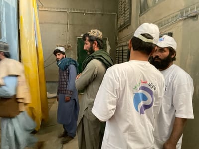 The image depicts an indoor scene within a functional building in Shuyene Vosta, Afghanistan, featuring five adult males. On the far right, two men are observed from behind and the side. Both wear white t-shirts featuring a multi-colored logo (blue, purple, red) and Dari script on the back, along with white baseball caps. The man closer to the right frame edge has a dark beard. To their left, a man with a dark beard, a patterned cap, and a dark green scarf draped over his traditional light-colored attire stands with his arms crossed. Adjacent to him, another man with a dark beard and white cap wears a dark blue scarf and a blue traditional garment with a patterned vest, looking directly forward. On the far left, a blurred figure in light-colored traditional clothing and a dark cap is partially visible, indicating motion. A prominent yellow, vertical fabric duct or curtain is positioned on the left side of the frame. The environment features light-colored walls with visible electrical conduits and wiring near the ceiling, and a concrete floor.