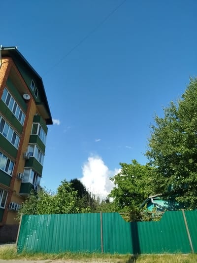 The image depicts an exterior scene in Olevsk, Ukraine, on a clear day with a bright blue sky. In the foreground, a green corrugated metal fence runs horizontally across the frame. Beyond the fence, lush green trees and foliage are visible, obscuring part of a small turquoise building with a visible roofline. To the left, a multi-story apartment building with yellow and green siding, featuring numerous windows and balconies, stands prominently. The building's architecture suggests it is a residential structure. The overall environment appears to be a mixed residential and natural area within the city. There are no people or animals visible in the scene.