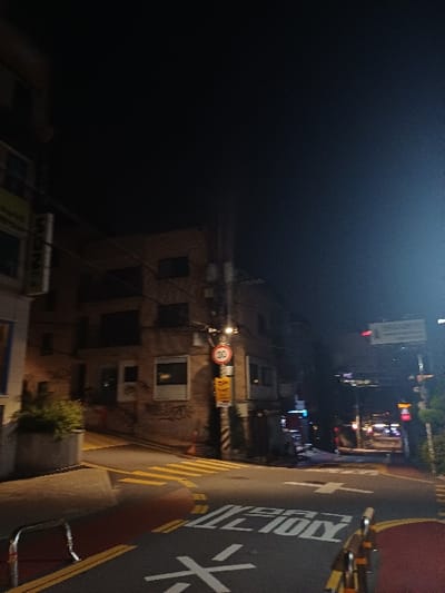 The image depicts an urban street intersection at night in 마포구, South Korea. The scene is illuminated by artificial streetlights, casting shadows. On the left, a multi-story building with a partially visible vertical sign reading "SSN" is present. To its right, another multi-story brick or stone building features multiple windows and visible graffiti on its lower facade.
The asphalt road exhibits clear markings: a yellow zebra crosswalk, a large white arrow indicating a straight direction, and Korean text "어린이 보호구역" (Children's Protection Zone) painted in white, followed by a white "30" speed limit marking. Sections of the road are painted red, consistent with a Children's Protection Zone.
A utility pole stands near the center-right, bearing a circular red "30" speed limit sign and several yellow rectangular signs beneath it, one of which denotes a Children's Protection Zone. Orange traffic cones are placed near the base of this pole. In the distance to the right, an overhead white sign is visible, along with the lights of vehicles. Metal barriers or guardrails line the right side of the roadway. A concrete planter with green foliage is situated on the left sidewalk. No people or active interactions are visible in the scene.
