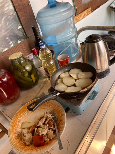 The image depicts a cluttered indoor space, possibly a kitchen, filled with various objects and food items.
A stainless steel electric kettle stands behind a black frying pan filled with sliced potatoes. To the left of the kettle is a large, blue water jug.
Beneath the pan and jug are several glass bottles, including ones containing cooking oil, wine, and homemade pickles in a large jar.
To the right of the pan is a plate of cooked food, including meat and potatoes. A fork rests on the plate.
The environment appears disorganized with a mix of textures and materials in the background.
The image was taken in the city of Urussu, Russia.