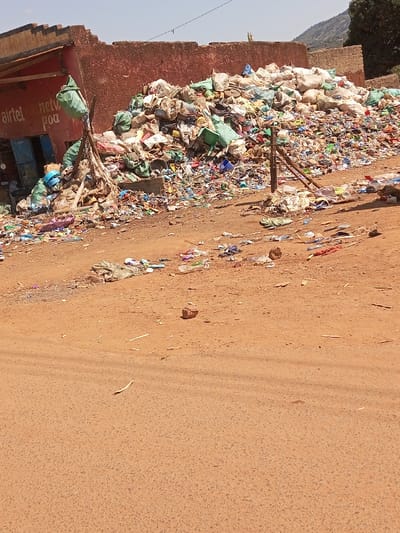 The image captures a static outdoor scene in Mukuyuni, Kenya, dominated by a substantial accumulation of refuse. The foreground presents a wide, unpaved reddish-brown dirt road, exhibiting faint linear depressions and scattered debris including small plastic bottles, fragments of plastic bags, and wood chips. A single reddish-brown brick is visible on the road surface near the bottom center.
To the mid-left, a partial view of a single-story building is visible. Its facade is painted red with white text, partially obscured, reading "airtel netw poa." The building features a blue-colored doorway and a small, square window. Green plastic bags are suspended from a wooden support structure that leans from this building towards the adjacent refuse pile.
The central and mid-right portions of the image are occupied by a large, irregularly shaped mound of garbage. This pile consists of a dense mixture of various discarded materials, prominently featuring numerous plastic bags (many white and green), plastic bottles, and other indistinguishable refuse items. Two rough wooden posts are positioned within or immediately adjacent to the garbage pile on the right side.
In the background, behind the refuse mound and the building, a reddish-brown wall or structure is visible. Beyond this, green hills or mountains rise under a clear, pale blue sky, with some distant vegetation on the far right. No people or animals are present in the depicted scene.