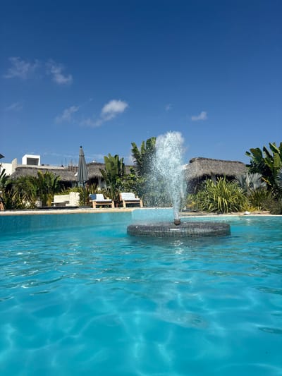 The image captures a vibrant outdoor scene next to a swimming pool in El Jícaro, Mexico. A water fountain feature is active, spraying water into the pool. The sky is clear and blue with some scattered clouds. Thatched-roof structures, possibly cabanas or buildings, are visible in the background amidst tropical vegetation, including palm trees and other greenery. Patio furniture, including lounge chairs and an umbrella, suggests a relaxation area near the pool. The pool water appears clear and turquoise, reflecting the sunlight.