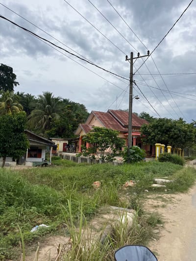 This media file captures an outdoor scene in Rantau Pauh, Indonesia, on a cloudy day. The foreground is dominated by lush green grass and foliage, with some scattered debris, suggesting a slightly unkempt area. A dirt road or pathway is visible on the right, also lined with grass. A part of a motorcycle's rearview mirror is visible on the far right.

In the middle ground, there is a residential area with several houses. One house, with a reddish-brown tiled roof, is a prominent feature, with yellow and white accents around its columns and windows. It appears to be a multi-story structure. Behind and around it, dense green trees and palm trees create a tropical backdrop. Another smaller building, partially visible below the larger house, has a dark-colored roof and some windows.

A significant element of the scene is a utility pole with multiple electrical wires crisscrossing the sky. The wires are a prominent visual line in the image, extending across the frame and connecting to the pole. The sky itself is overcast, with dramatic grey clouds, indicating a possibly humid or pre-rainy atmosphere. The lighting is diffused, typical of a cloudy day, with no strong shadows.

The overall impression is one of a rural or semi-rural residential area in a tropical climate, characterized by vegetation, modest housing, and essential infrastructure like power lines. There is no visible text or indication of specific human activity beyond the presence of the houses and utility pole. The cloudy weather contributes to a somewhat subdued or tranquil mood.