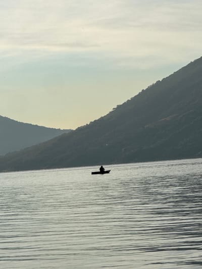 The image displays a solitary individual in a small, dark boat on the calm waters of Lake Atitlán, near San Pablo La Laguna, Guatemala. The boat, a narrow vessel, is positioned centrally in the mid-ground. A single person, depicted as a dark silhouette, is seated within the boat, appearing to wear a wide-brimmed hat. The water exhibits subtle, horizontal ripples across its surface. In the background, two prominent mountain slopes rise from the lake. The closer slope on the right is densely covered with vegetation and appears darker, while the more distant slope on the left is lighter and hazy. The sky is a pale, diffused blue-grey, indicating either dawn or dusk.